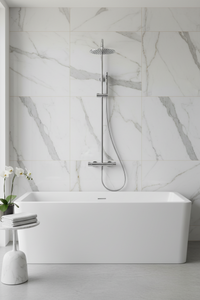 A modern freestanding rectangular bathtub in white, set against a large white marble tiled wall with a chrome rain shower head and hand shower mounted above it. A small white side table with folded towels and a flower arrangement sits to the left.